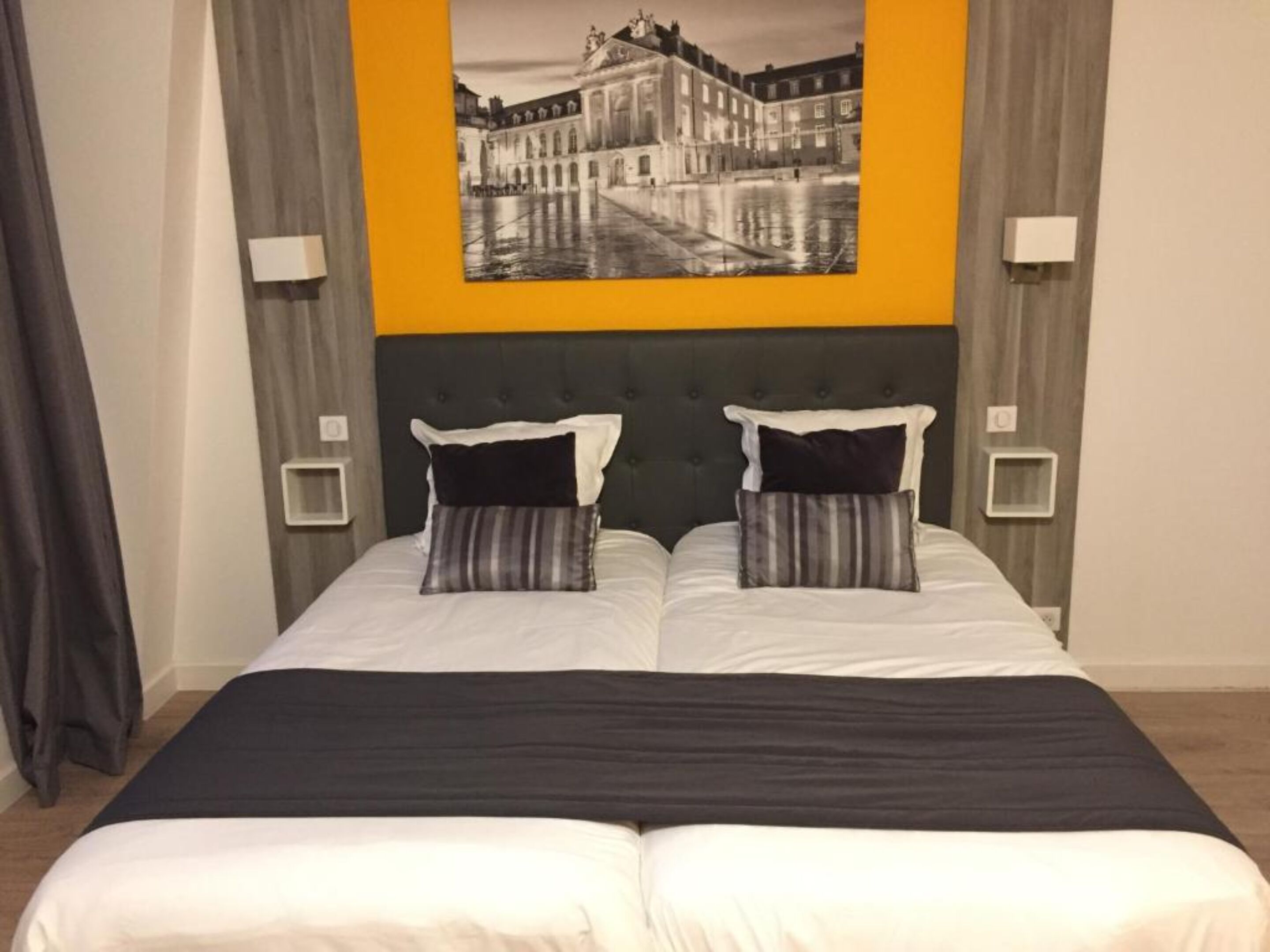 Una cama doble con dos almohadas, un cabecero oscuro y una amplia pintura en blanco y negro de un paisaje urbano.