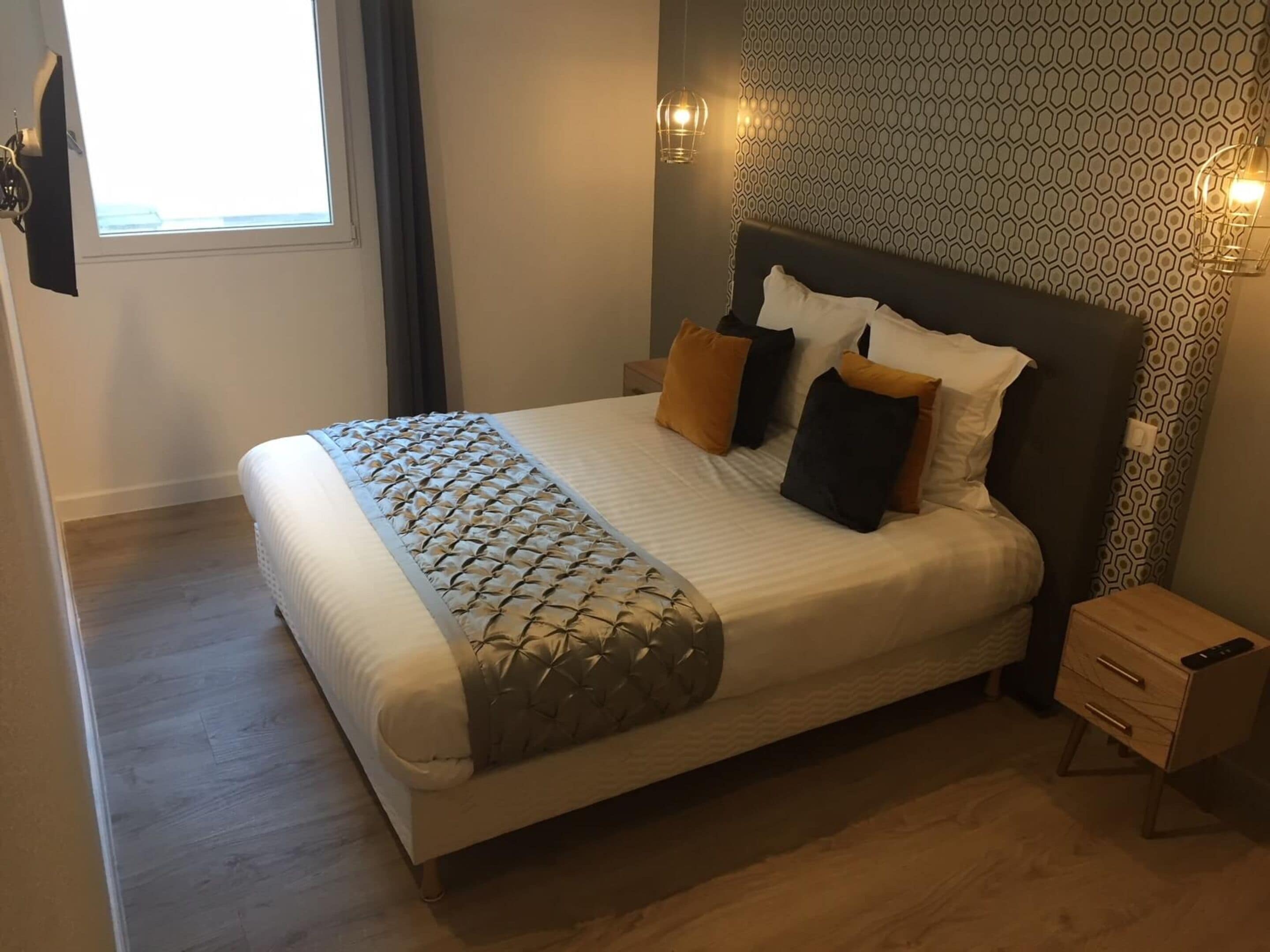 A neatly made bed with a patterned bedspread, two bedside tables with drawers, a wall with a geometric pattern, and a window with curtains.