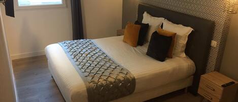 A neatly made bed with a patterned bedspread, two bedside tables with drawers, a wall with a geometric pattern, and a window with curtains.