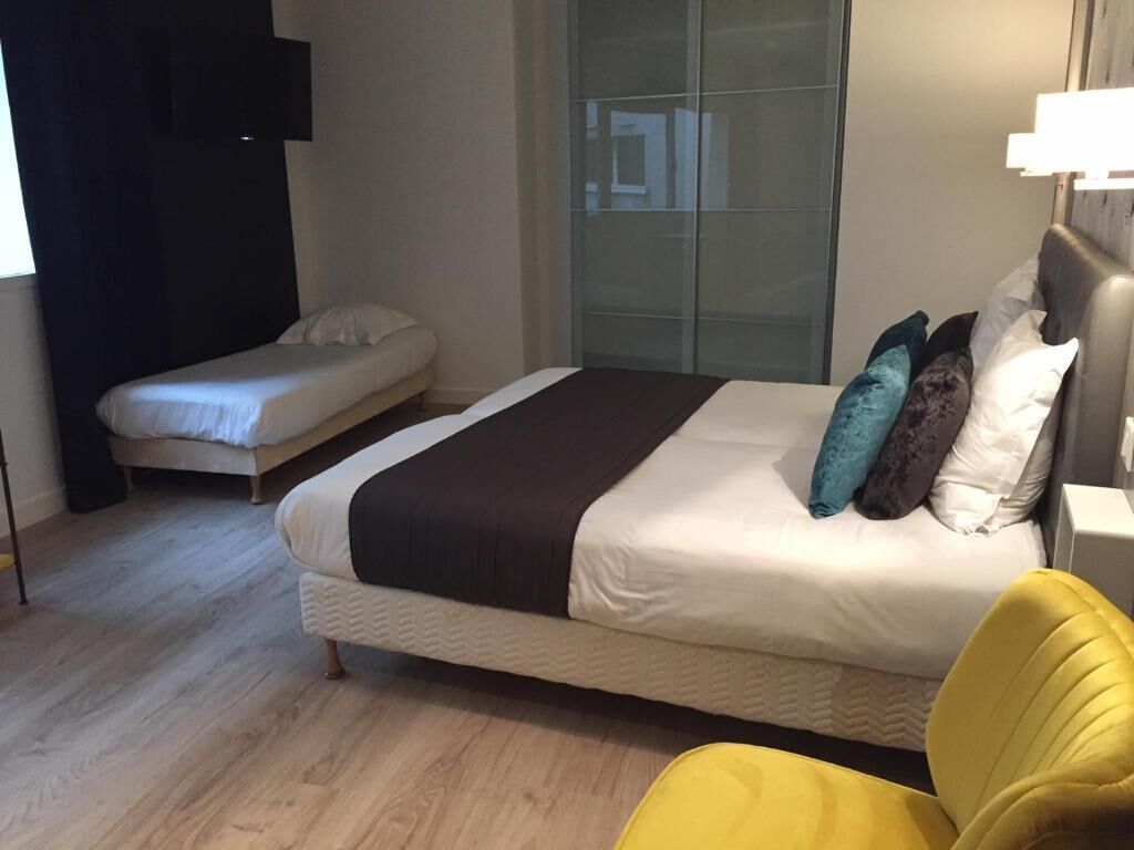 Une chambre avec un grand lit, un plus petit lit, une chaise jaune et une cloison en verre.