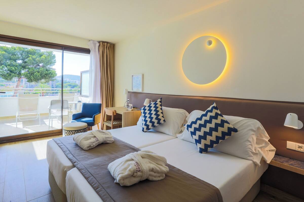 A hotel room with a bed, two pillows, a bedside table with a lamp, a chair, and a view of the outdoors.