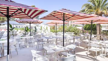 Un área de comedor al aire libre con mesas y sillas blancas, sombrillas a rayas rojas y blancas, y palmeras al fondo.