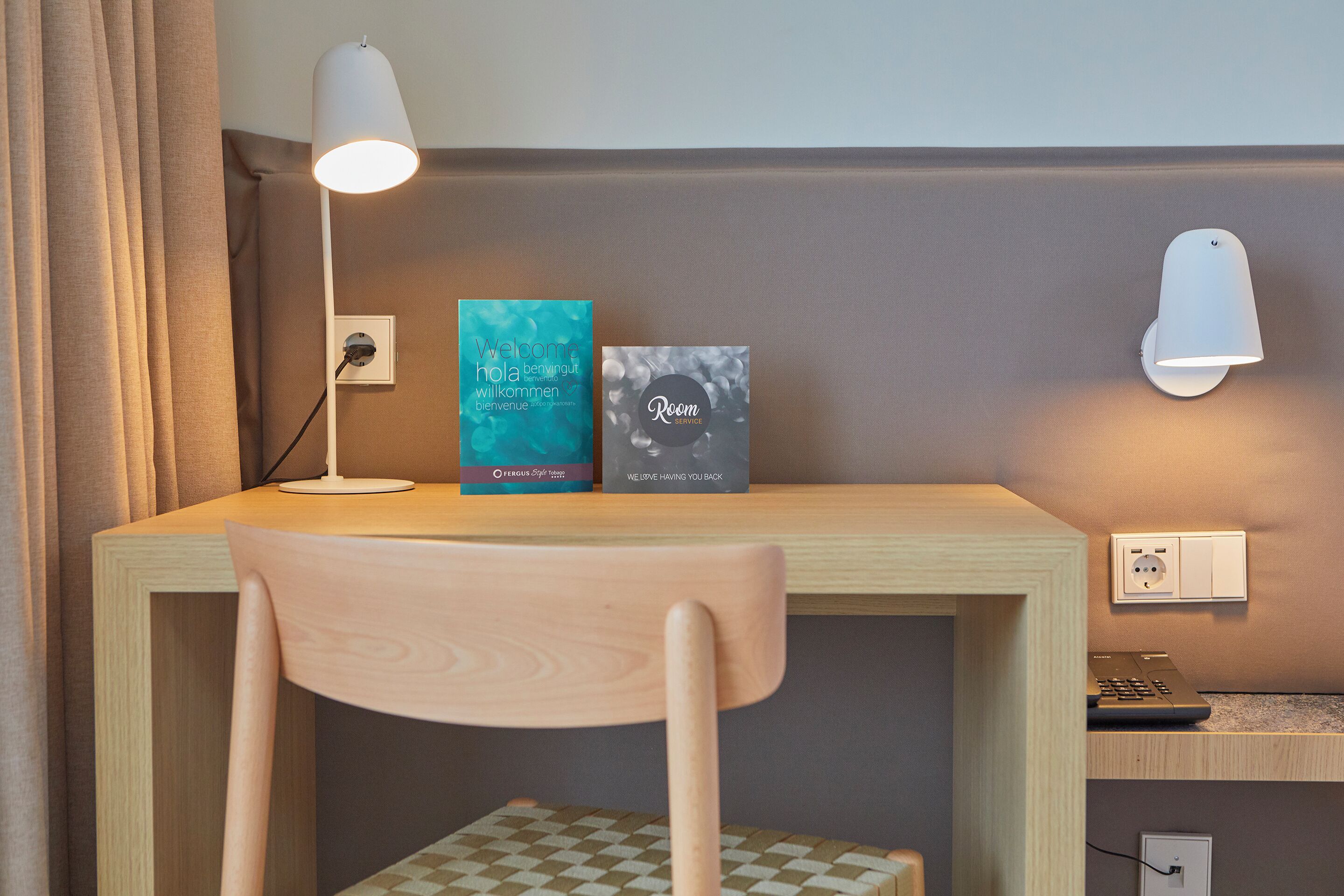 A desk with a chair, two books, a lamp, and a wall-mounted light fixture.