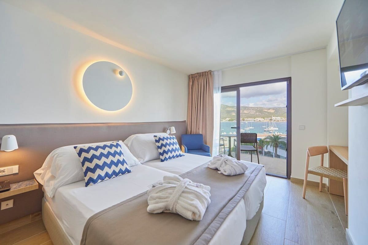 A hotel room with a large bed, two pillows, a bedside table with a lamp, a chair, a TV, and a balcony with a view of the sea and boats.