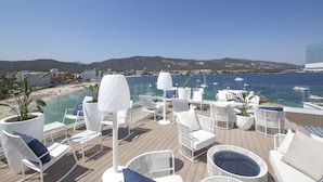 Terraza exterior iluminada por el sol con sillones blancos y una bañera de hidromasaje, con vistas a una playa y al mar.