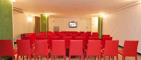 Un salón de conferencias con hileras de sillas rojas, una pantalla de televisión y una pared verde decorativa.