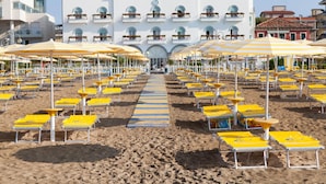A beach with rows of yellow sun loungers and umbrellas, a central blue-striped pathway, and a white building labeled "HOTEL MARINA" in the background. - Hotel Marina (Jesolo)