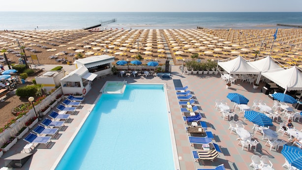 A pool area with sun loungers and umbrellas, a beach with umbrellas, and a pier in the background. - Hotel Marina (Jesolo)