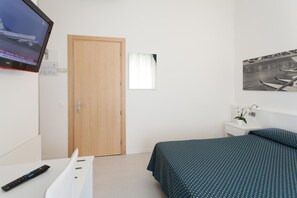 A bedroom with a bed, a TV mounted on the wall, a wooden door, a small window, and a nightstand with a plant. - Hotel Marina (Jesolo)
