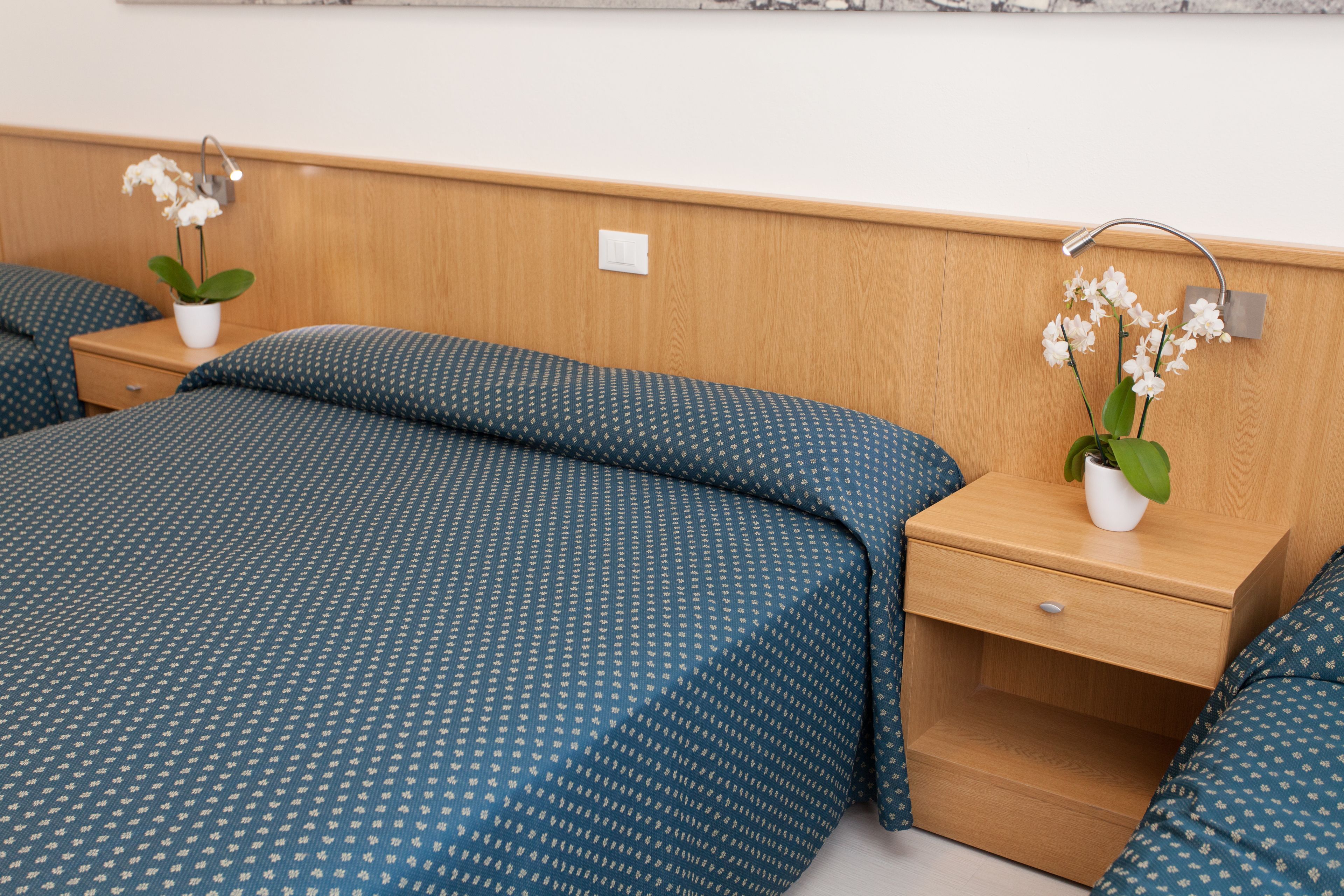 A hotel room with a wooden headboard, a single bed with blue patterned bedding, a nightstand with a potted orchid, and a bedside table with a lamp.