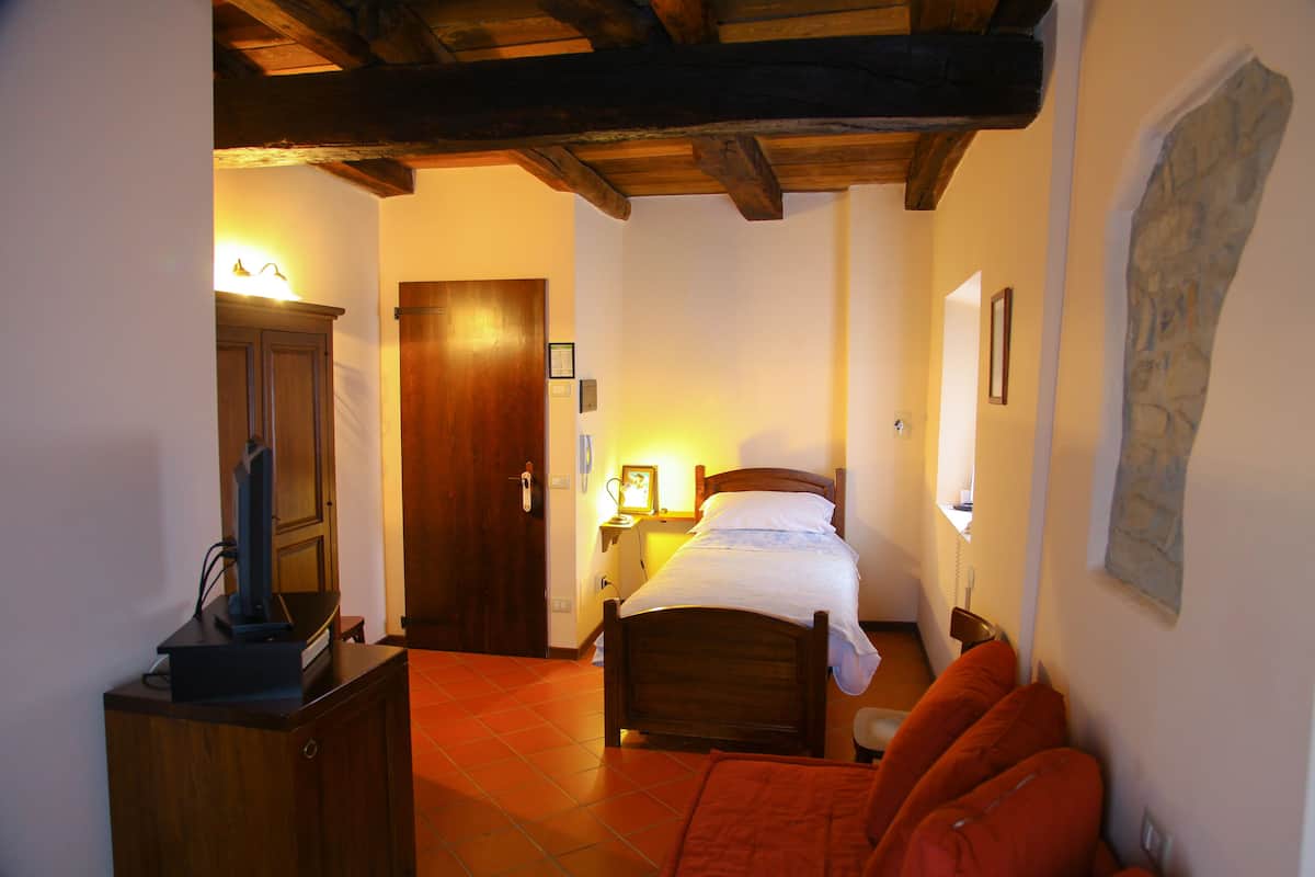 A bedroom with a wooden bed, a nightstand, a desk with a computer, a wardrobe, and a door leading to another room.