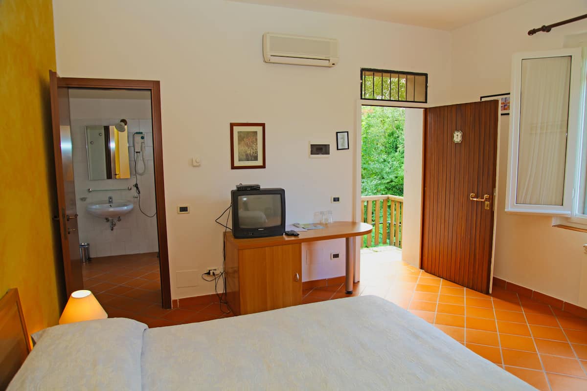 A bedroom with a bed, a wooden desk, a television, a mirror, and a bathroom visible through an open door.