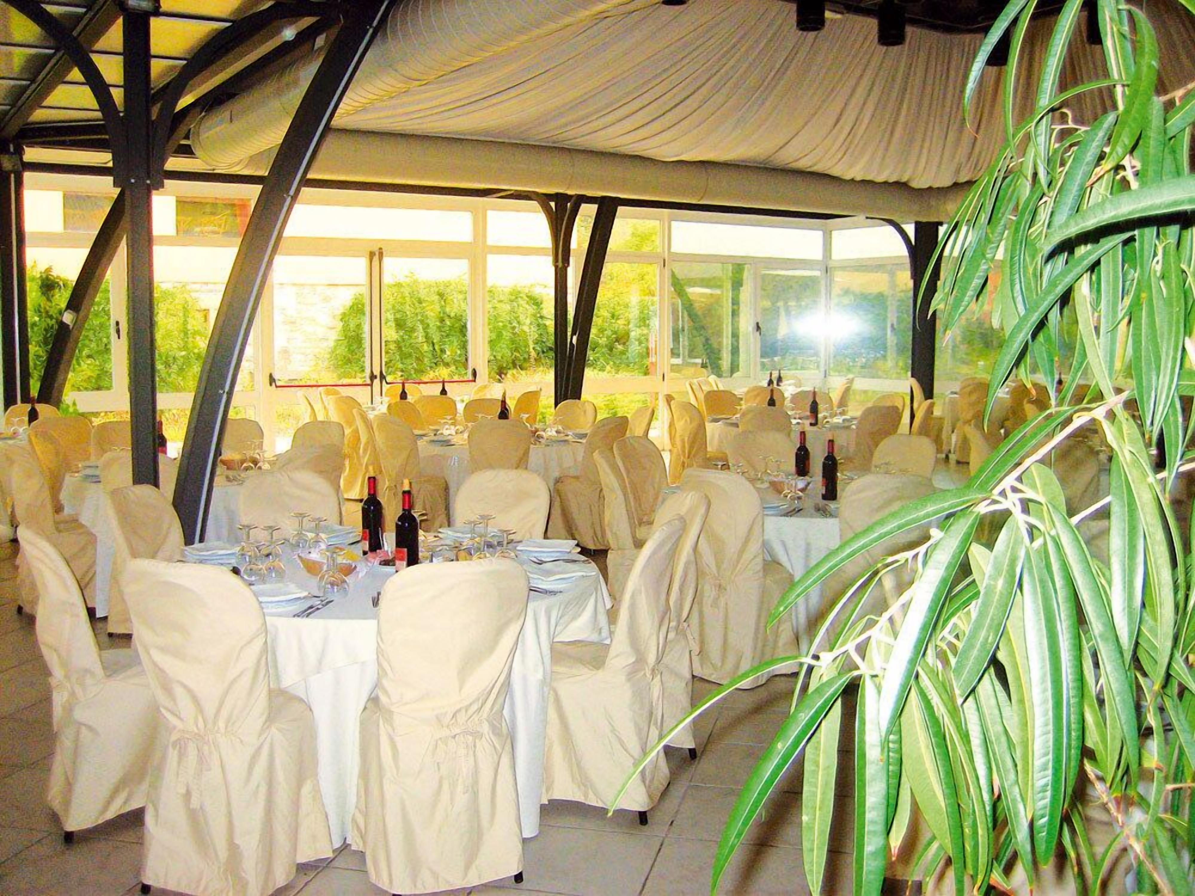 Une salle de banquet intérieure, avec des tables dressées pour le repas, recouvertes de nappes blanches et entourées de chaises, et de grandes fenêtres laissant entrer la lumière naturelle.
