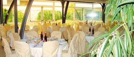 Une salle de banquet intérieure, avec des tables dressées pour le repas, recouvertes de nappes blanches et entourées de chaises, et de grandes fenêtres laissant entrer la lumière naturelle.