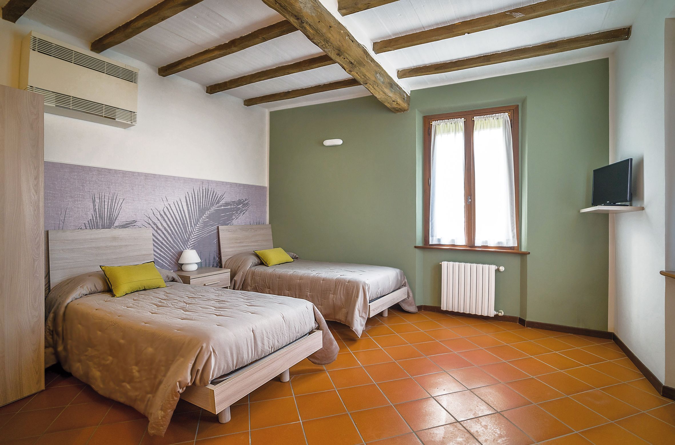Une chambre avec deux lits, une fenêtre avec des rideaux, un radiateur et un téléviseur posé sur une étagère.