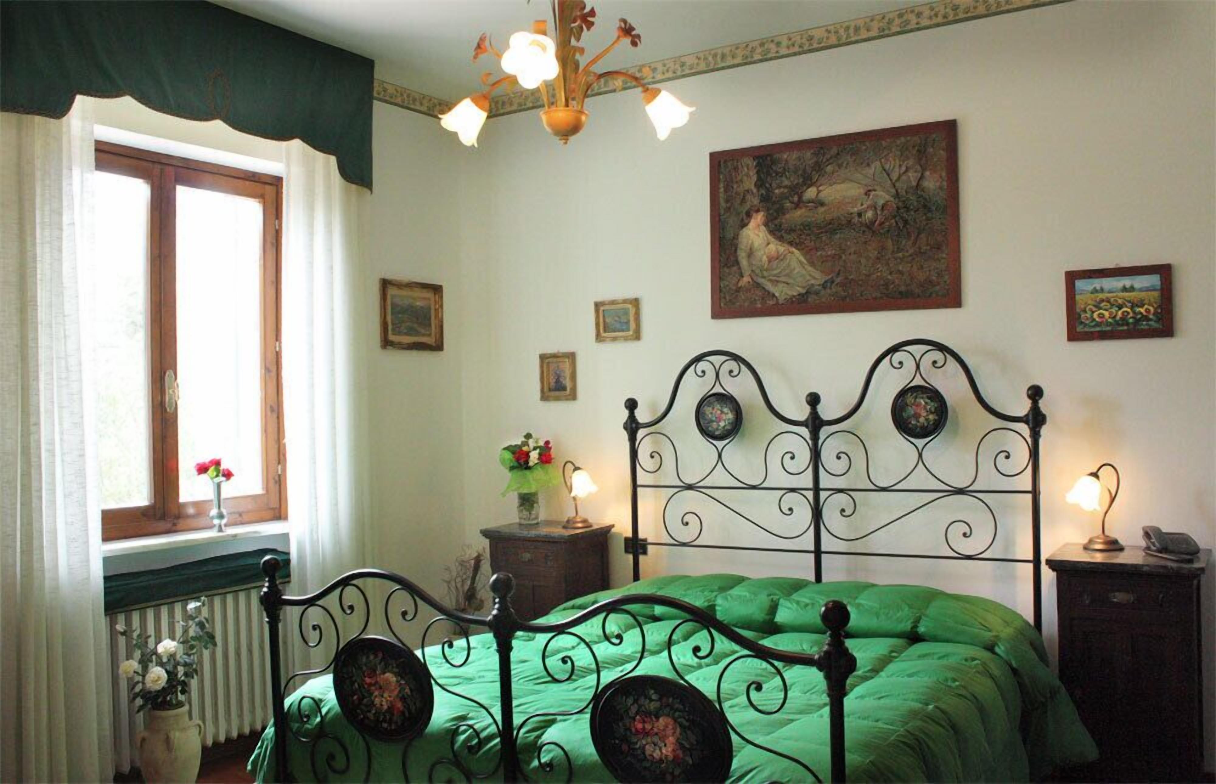 Un dormitorio con cama verde, cabecero de hierro decorativo, ventana con cortinas, radiador, un florero con flores y un cuadro en la pared.