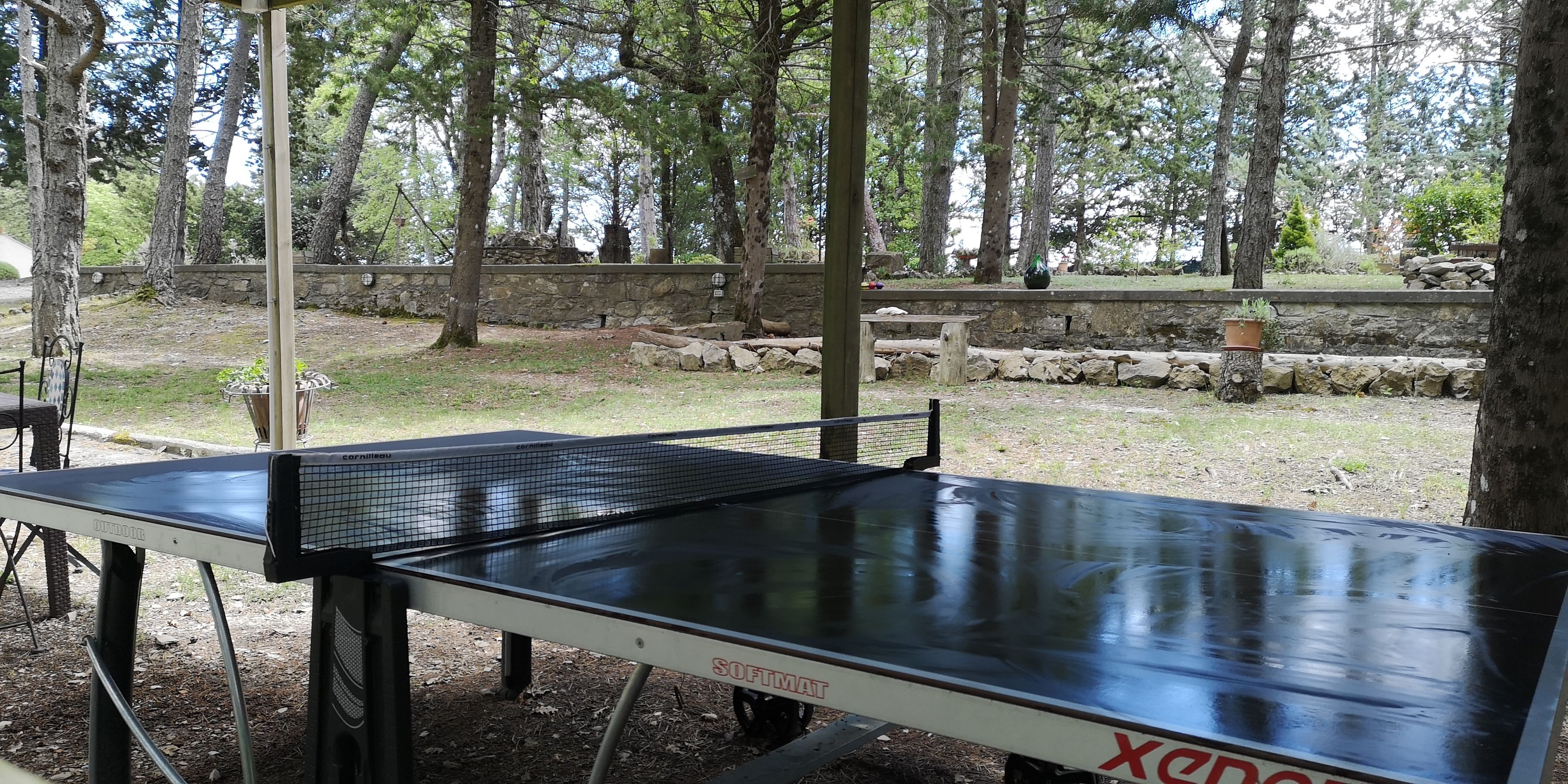 Mesa de ping pong al aire libre en zona boscosa con muros de piedra y árboles.