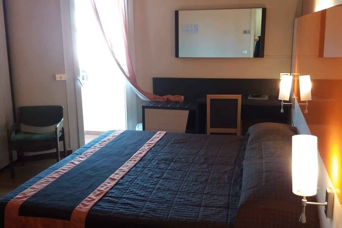 A hotel room with a bed, a desk, a chair, a bedside table with a phone, and a window with curtains.