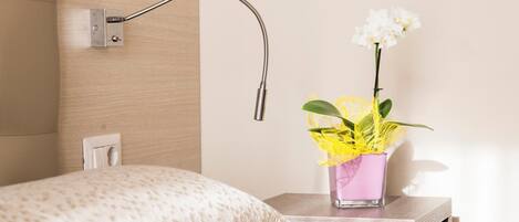 A neatly arranged hotel room with a bed, a nightstand with a vase of flowers, and a wall-mounted lamp.