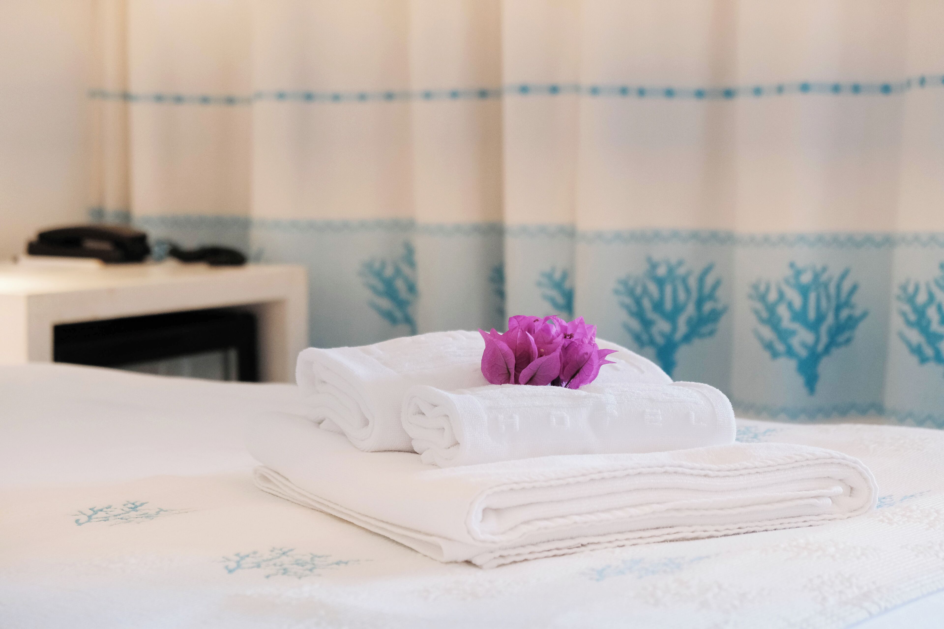 A neatly made bed with folded white towels and a purple flower on top.