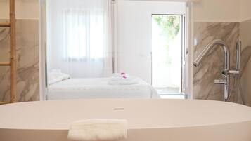 A modern bathroom with a freestanding bathtub, a shower area, and a visible bedroom with a bed and a window.