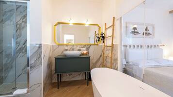 Un baño moderno con ducha de mármol, un lavabo con encimera oscura, un espejo grande con marco dorado y una cama con cabecero blanco.