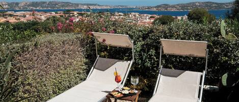 Two sun loungers with a table and drinks, overlooking a coastal town and hills.