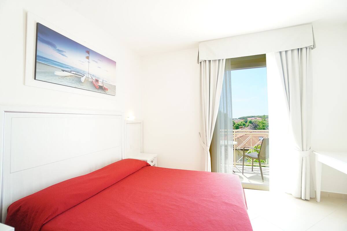 Une chambre d’hôtel avec un lit, un tableau représentant une scène de plage, un balcon avec une table et une chaise, et une vue sur la campagne.