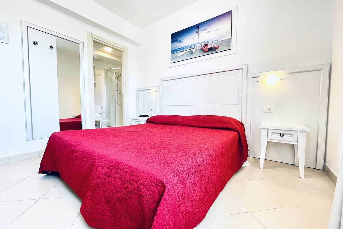 Une chambre à coucher avec une couvre-lit rouge, une table de chevet blanche, une commode, un tableau représentant une scène de plage et une porte donnant sur une salle de bain.