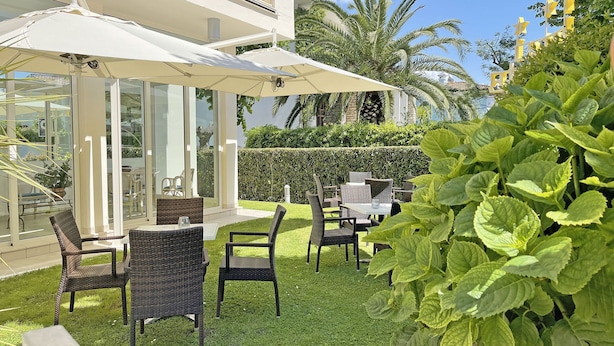 An outdoor patio with wicker furniture and white umbrellas, surrounded by lush greenery and palm trees. - Hotel Pigalle (Forte dei Marmi)