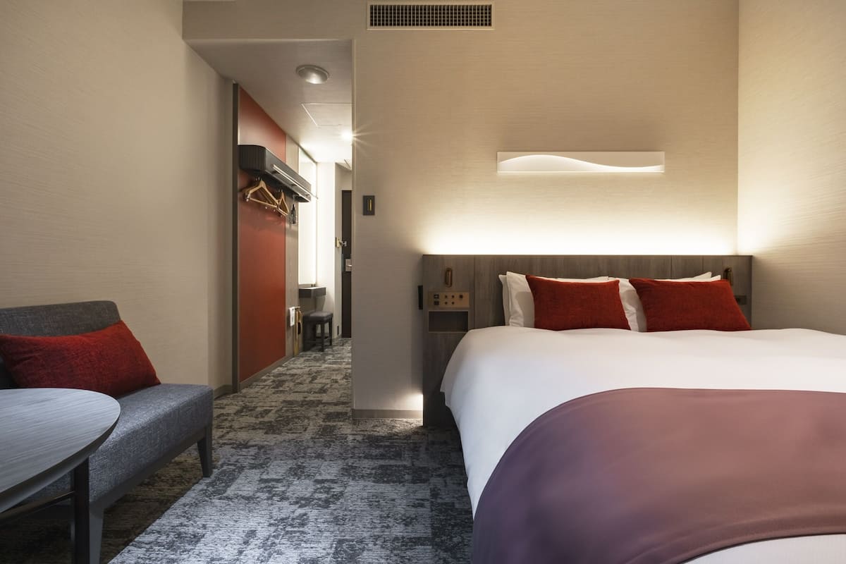 A hotel room with a bed, a desk, and a chair. There is a wall-mounted light above the bed and a small table with a chair.