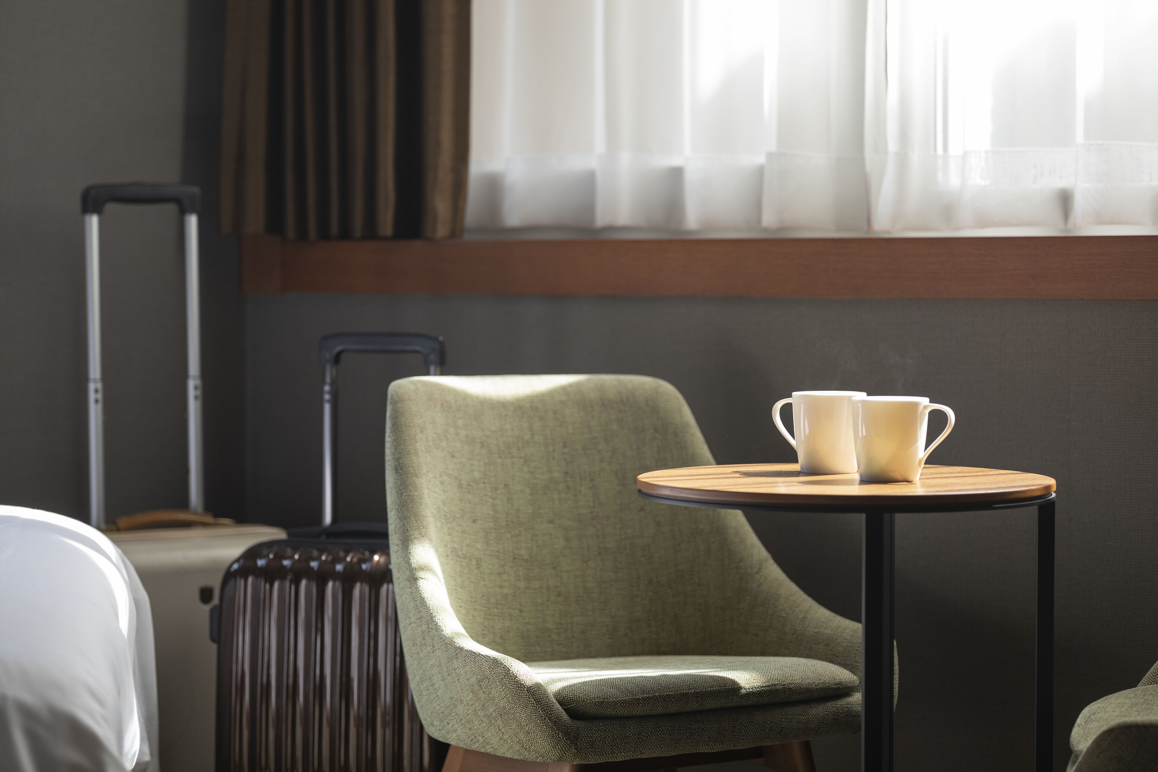 A room with a green upholstered chair, a small round table with two white cups, a suitcase, and a bed in the background.