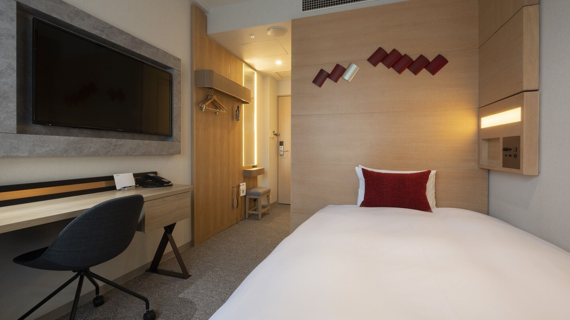 A hotel room with a bed, a desk, a chair, a TV, and a wall-mounted light fixture.