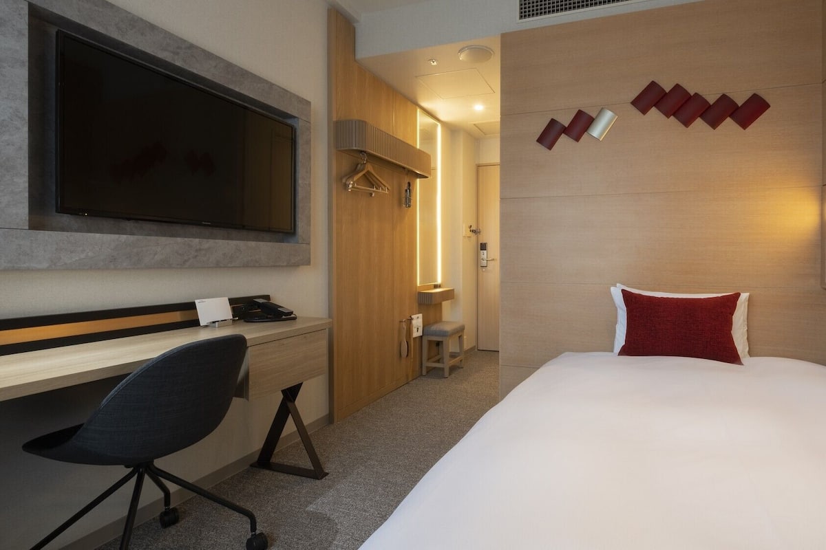 A hotel room with a bed, a desk, a chair, a TV, and a wall-mounted light fixture.