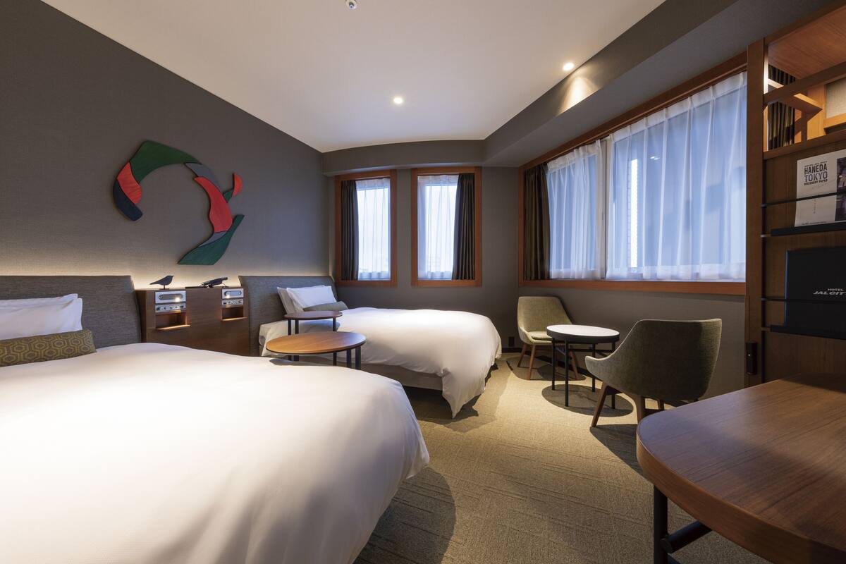 A hotel room with two beds, a desk, a chair, a small table, and a bookshelf.