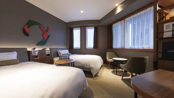 A hotel room with two beds, a desk, a chair, a small table, and a bookshelf.
