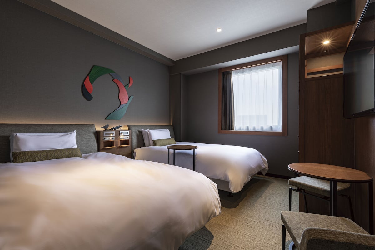 A hotel room with two beds, a small table, a chair, a TV, and a window with curtains.