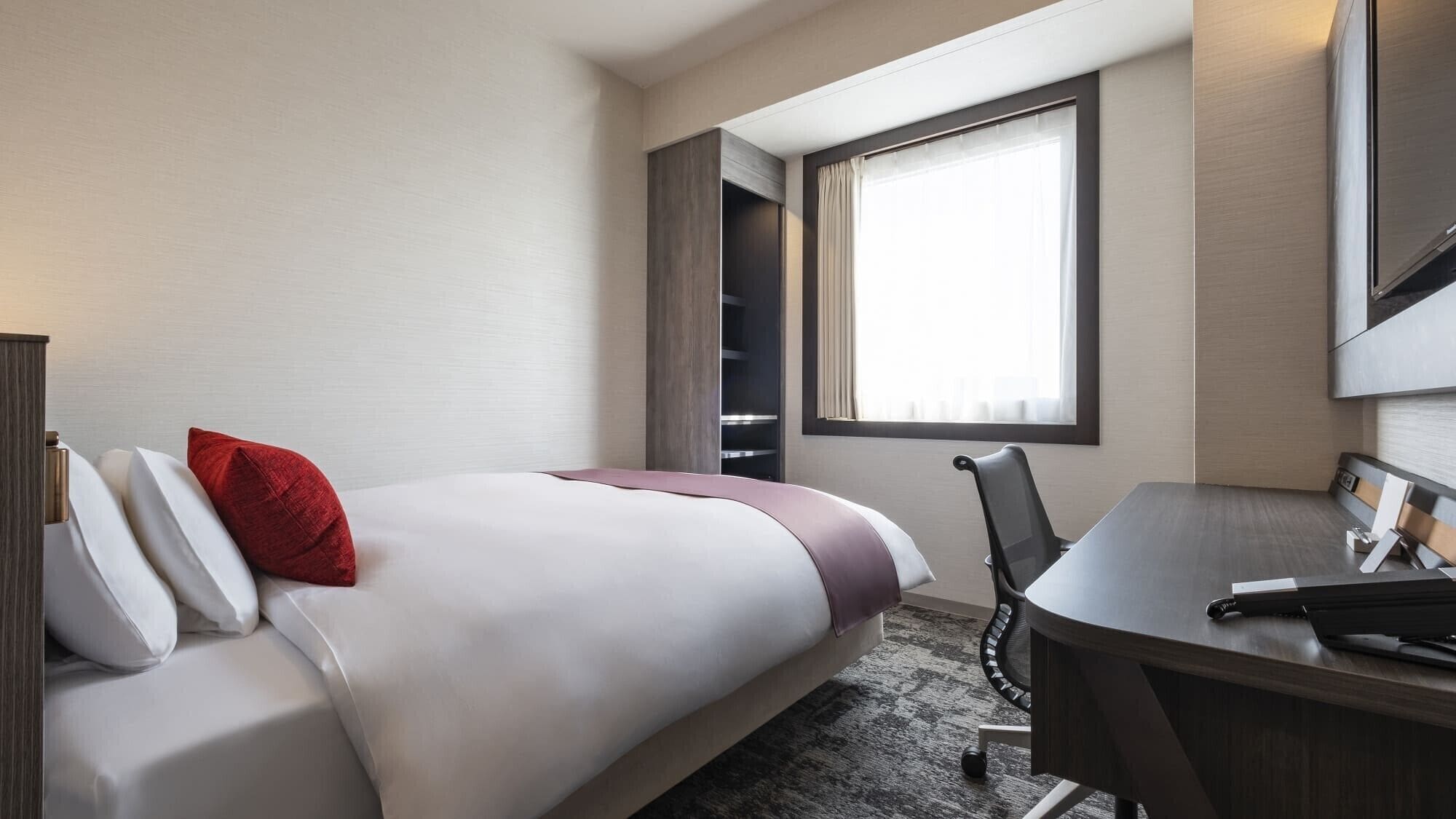 A hotel room with a bed, a desk, a chair, a window with curtains, and a wardrobe.