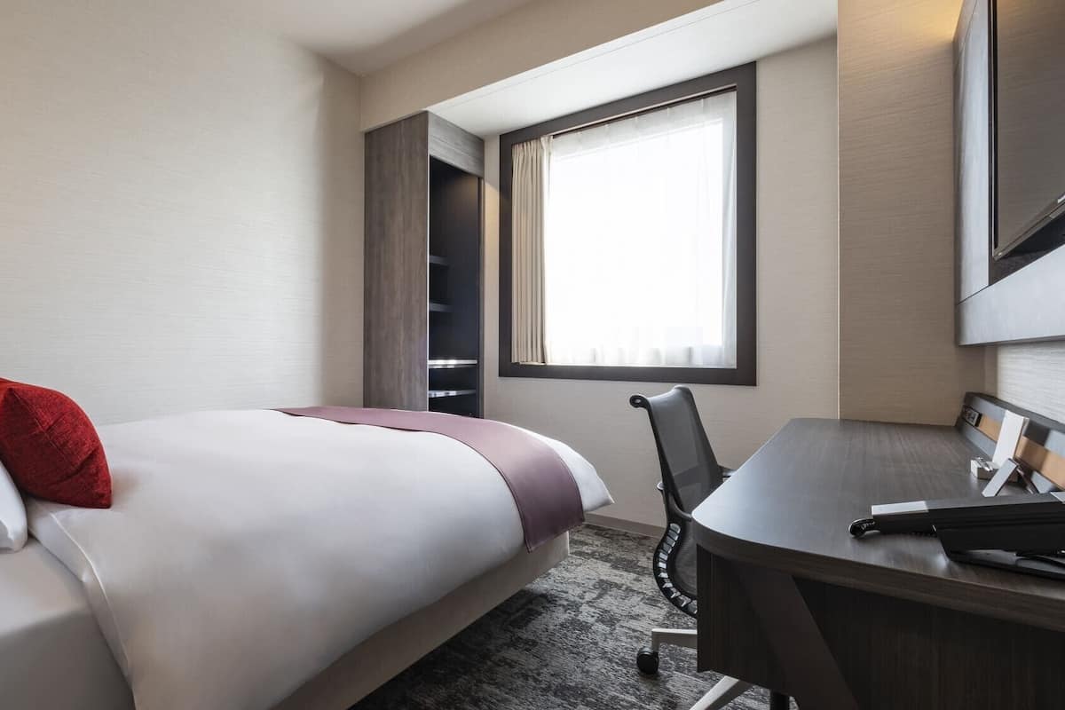 A hotel room with a bed, a desk, a chair, a window with curtains, and a wardrobe.