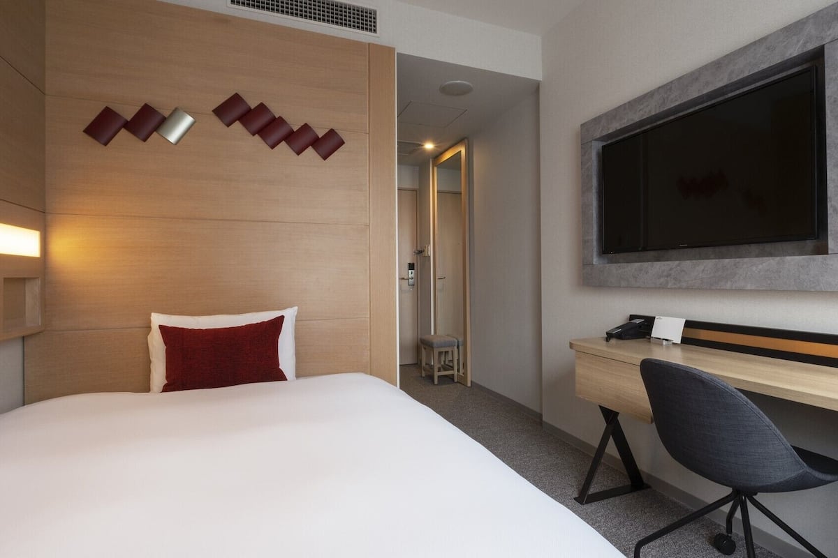 A hotel room with a bed, a desk, a chair, a TV, and a shower area.