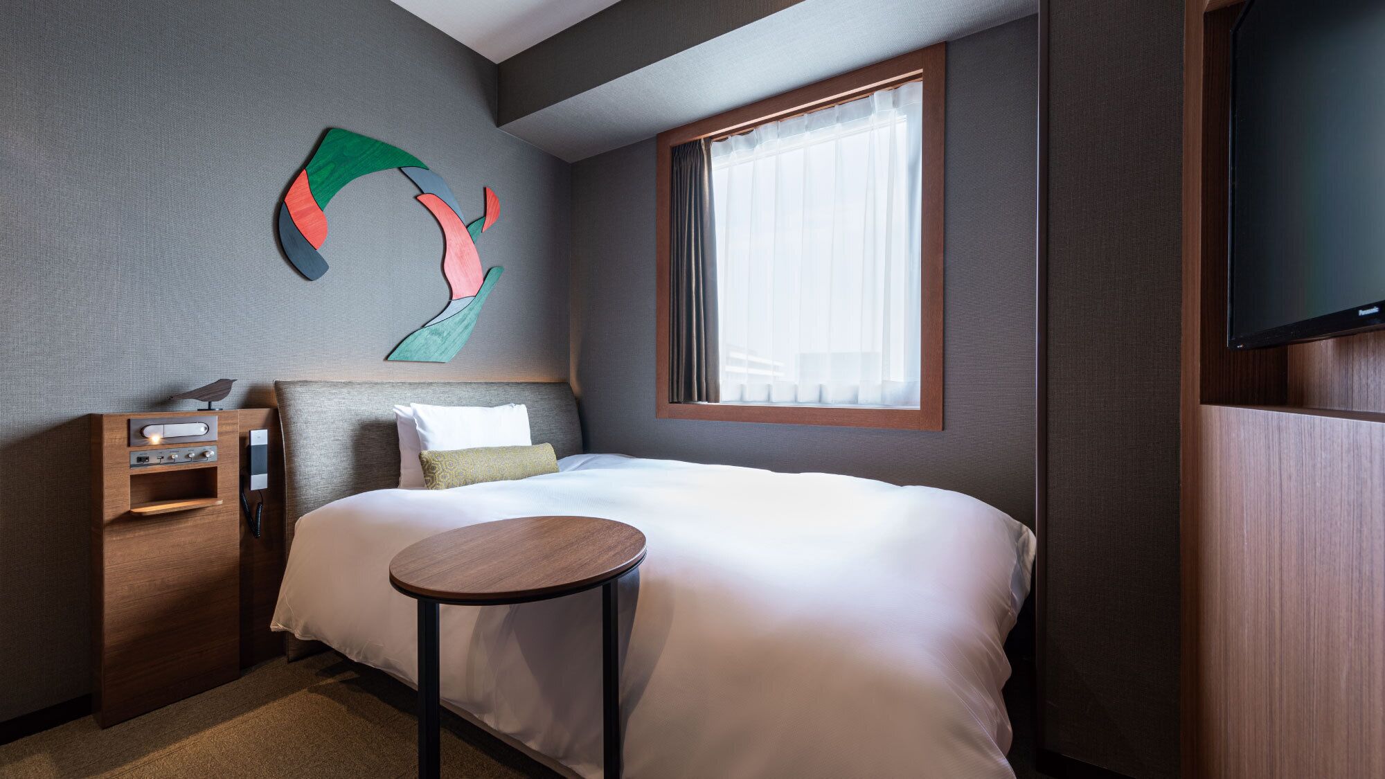 A hotel room with a bed, a bedside table, a wall-mounted TV, and a window with curtains.