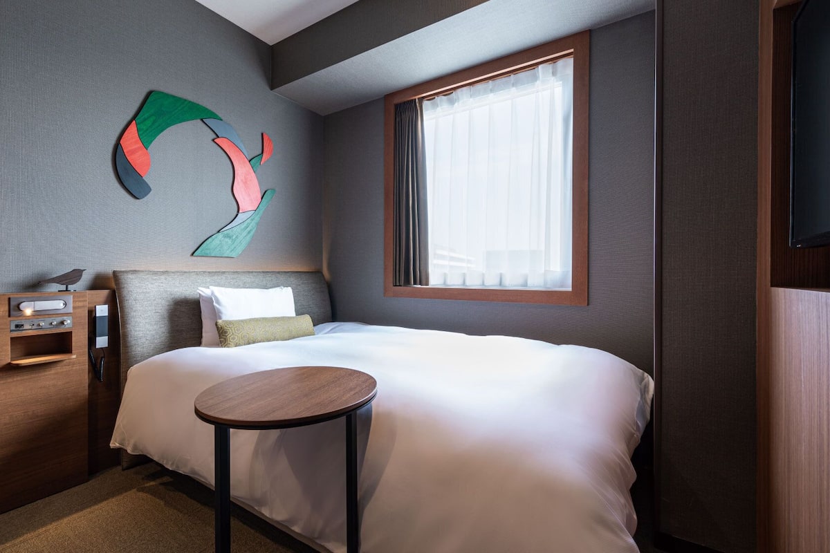 A hotel room with a bed, a bedside table, a wall-mounted TV, and a window with curtains.