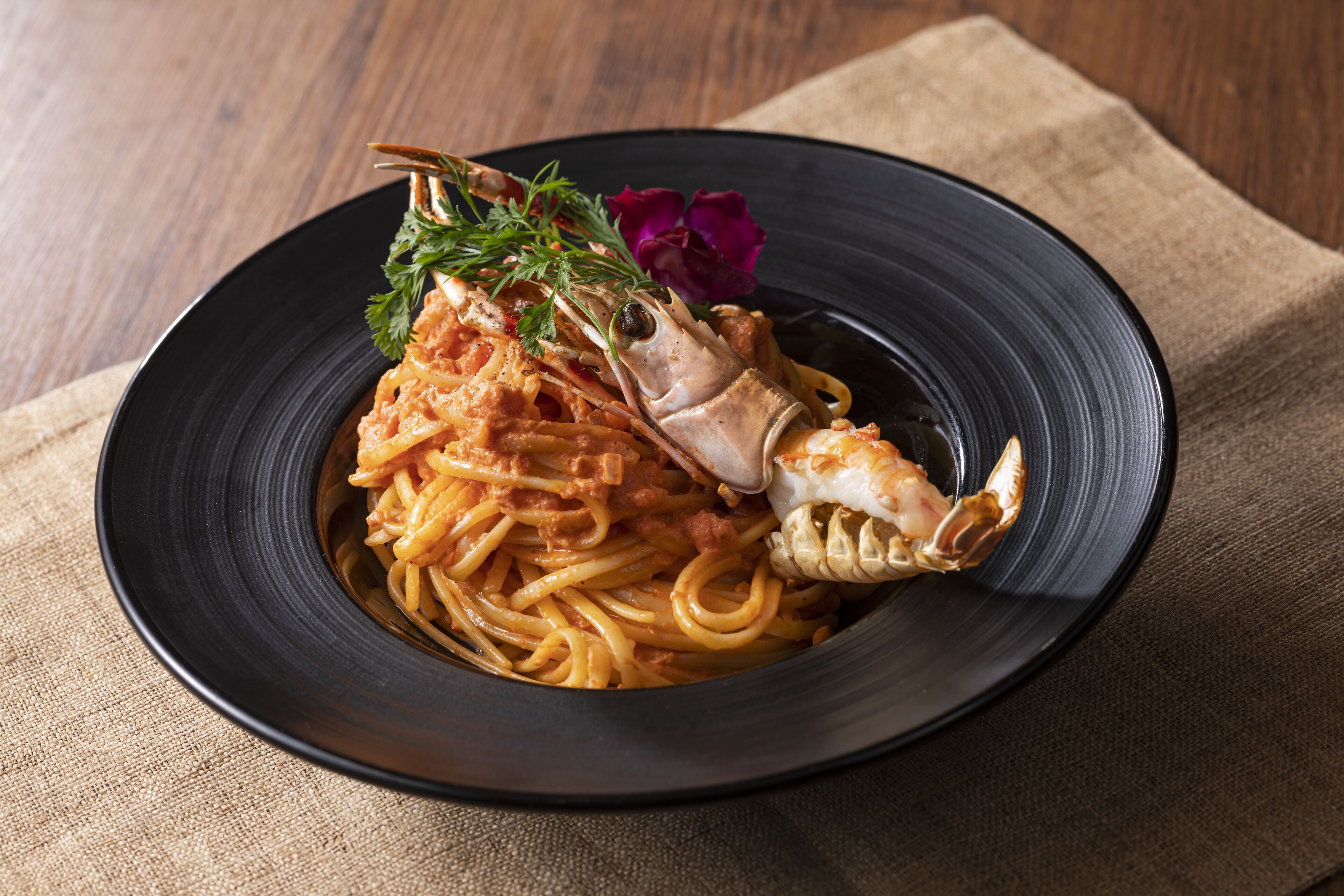A plate of spaghetti with a lobster tail and garnish on a black plate.