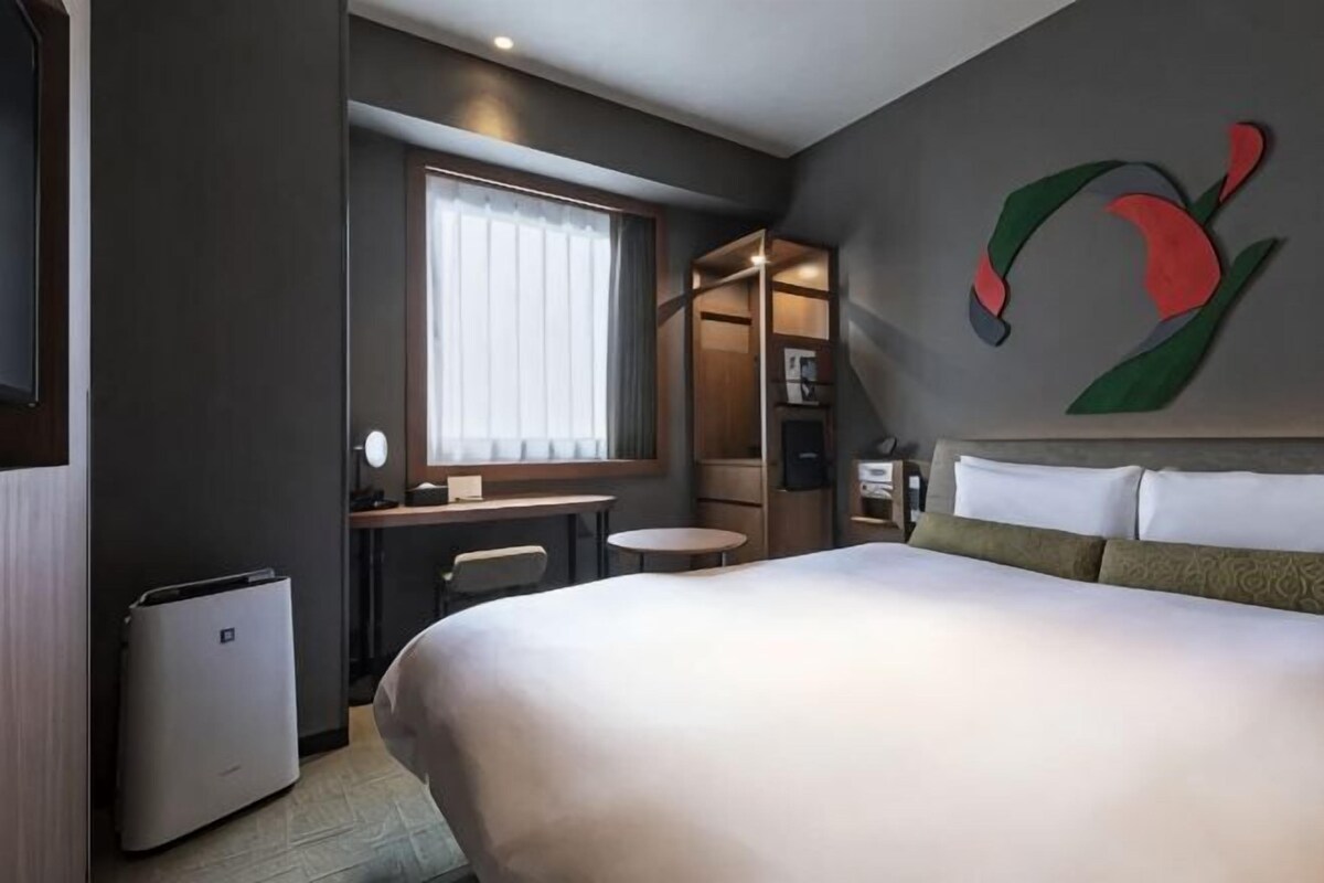 Habitación de hotel con una cama grande, un escritorio, una silla, una mesita, una ventana con persianas y una obra de arte en la pared.