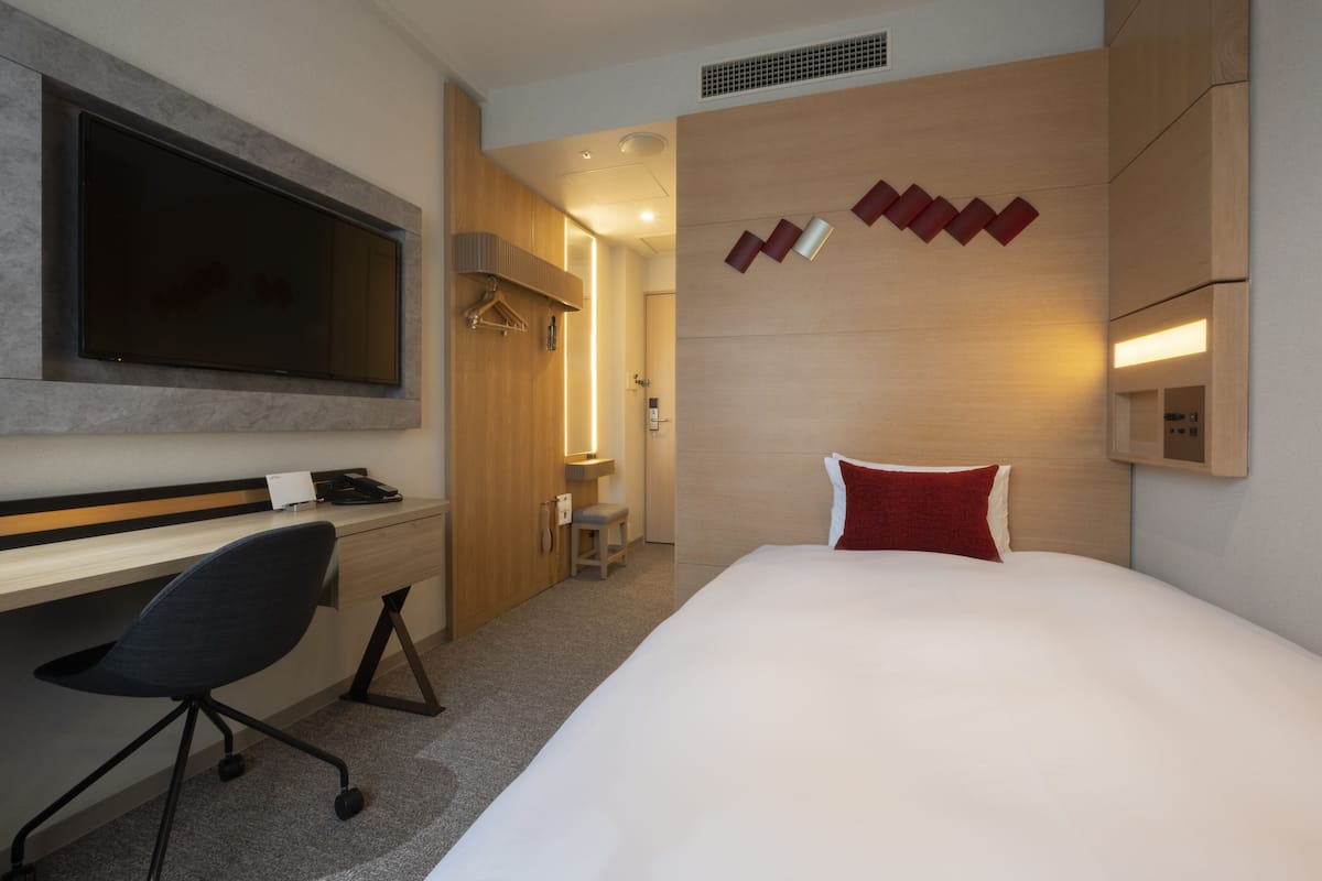 A hotel room with a large bed, a desk with a chair, a flat-screen TV mounted on the wall, and a small stool.