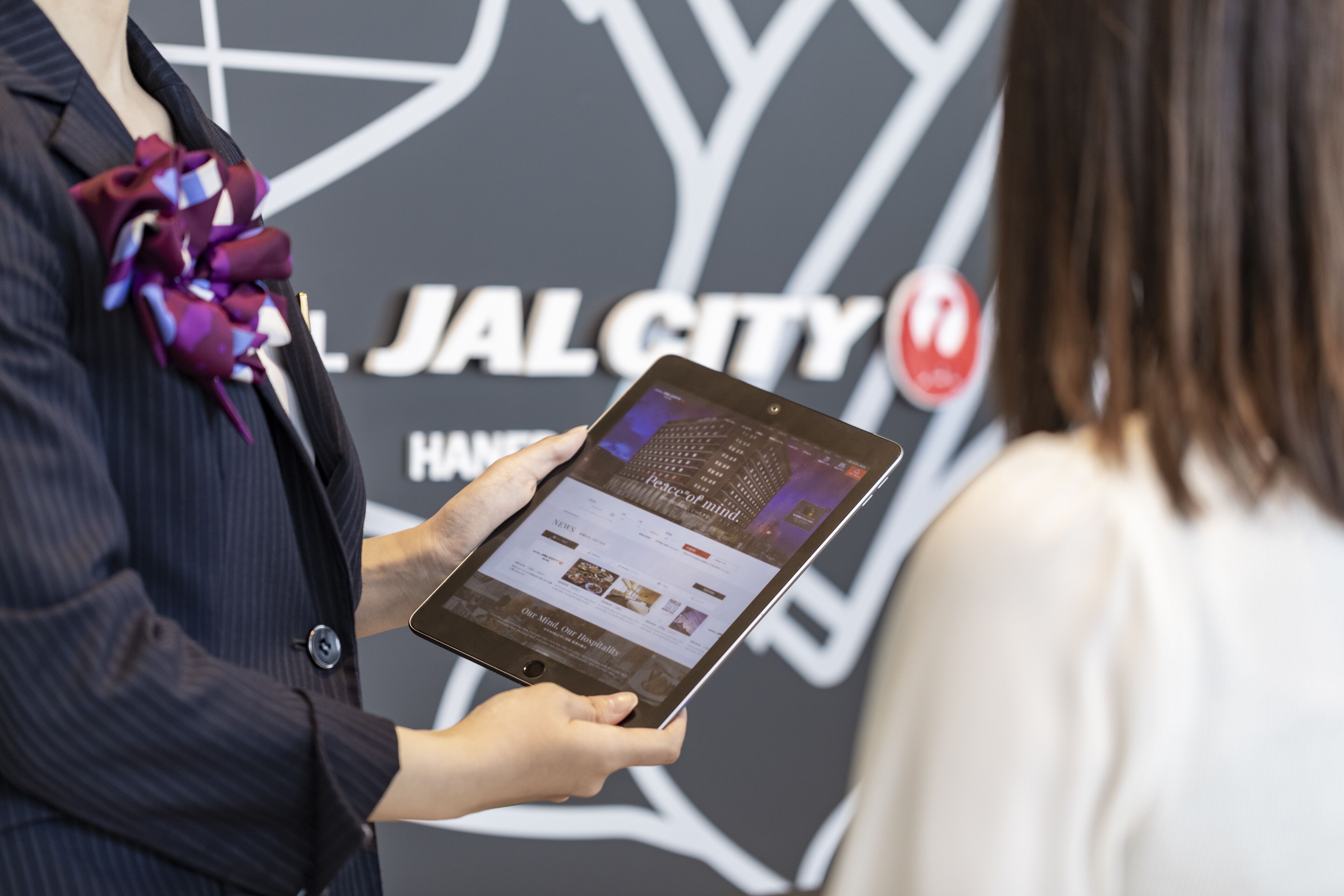 A person holding a tablet with a webpage on it, standing in front of a sign that reads "JAL CITY".