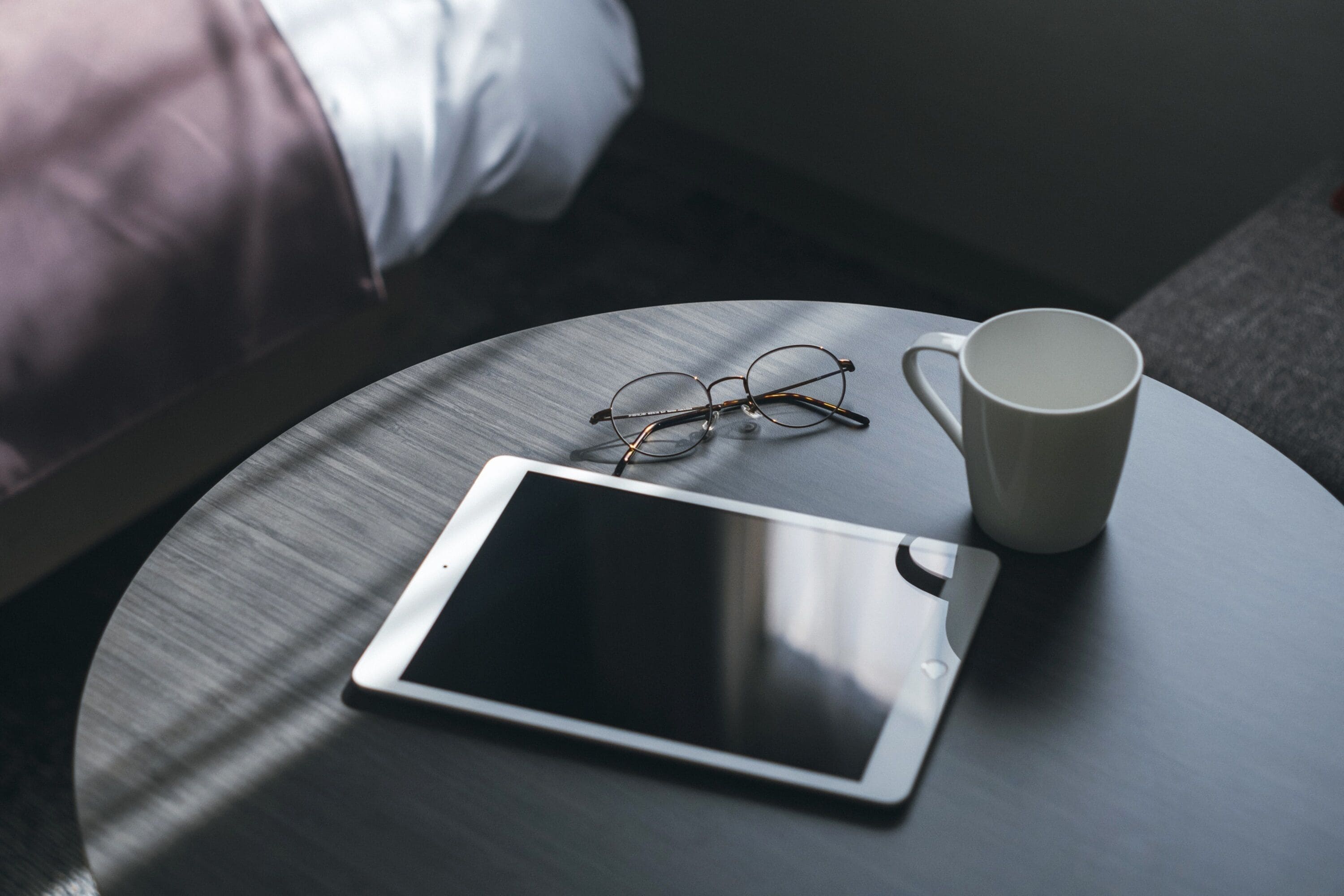Una tableta, un par de gafas y una taza blanca sobre una mesa redonda en una habitación.