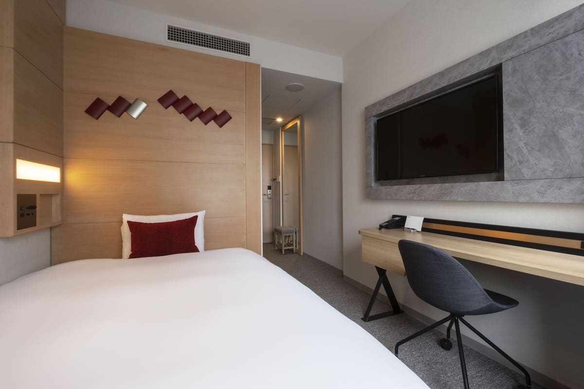 A hotel room with a large bed, a desk with a chair, a flat-screen TV, and a wall-mounted shelf with decorative items.