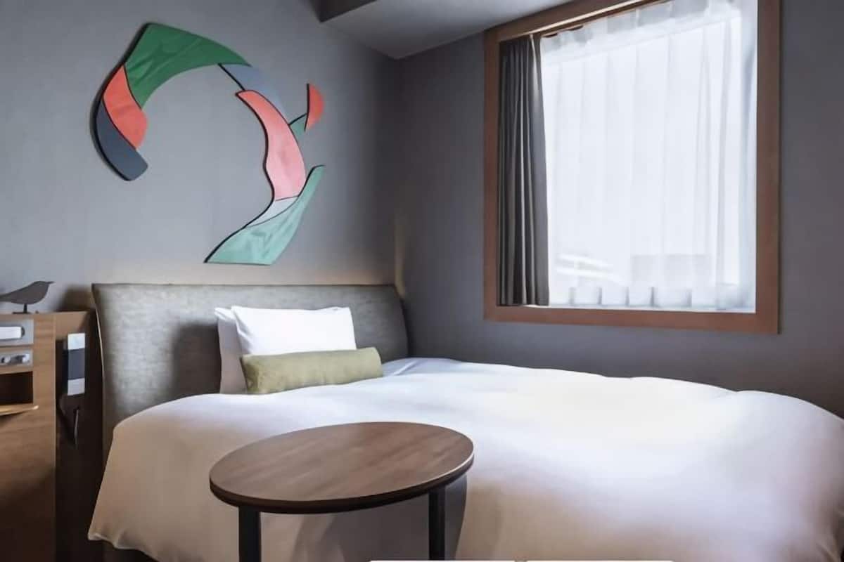 Una habitación de hotel con cama, una mesa redonda, una obra de arte en la pared y una ventana con cortinas.