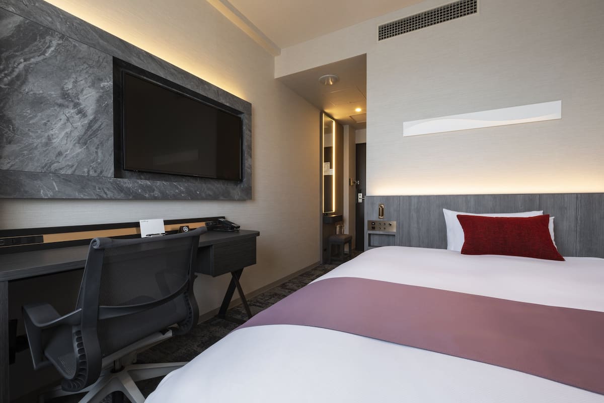 A hotel room with a bed, a desk, a chair, a TV mounted on the wall, and a red pillow.